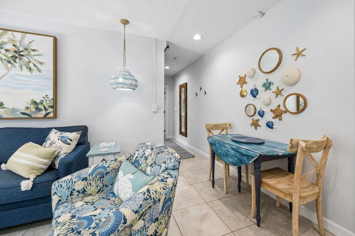 A living room with a sofa, a patterned armchair, a dining table with chairs, and decorative wall art including seashells and starfish.