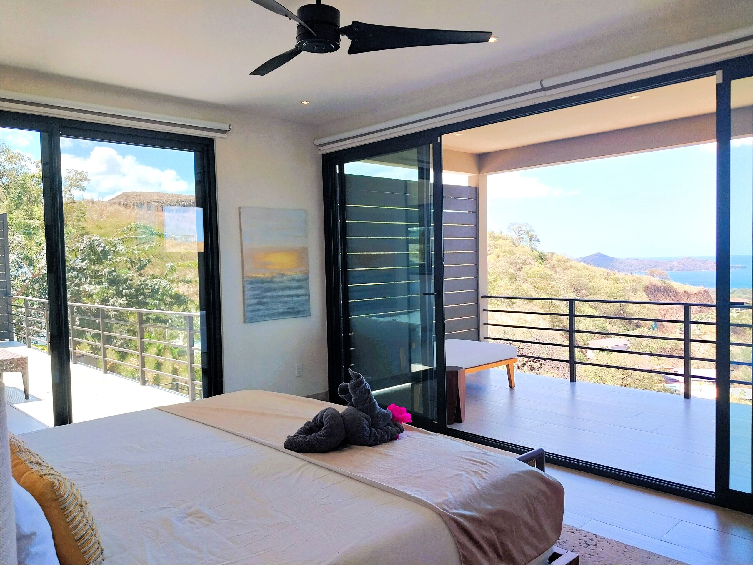 A bedroom with a bed, a ceiling fan, a large window, and a balcony with a view of the landscape.