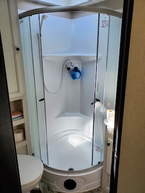 A compact, modern bathroom with a glass-enclosed shower, a toilet, and a shelf with toiletries. - Our Camper is perfect for a weekend away... (Sequim)