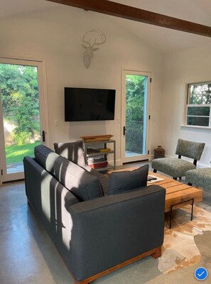 Relax in this stylish living room with a large sofa, flat-screen TV, and a glass door leading to a lush outdoor area.