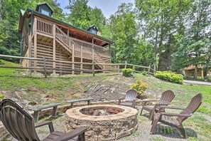 Una cabaña con un hogar de piedra y sillas Adirondack, rodeada por una terraza de madera y un muro de piedra.