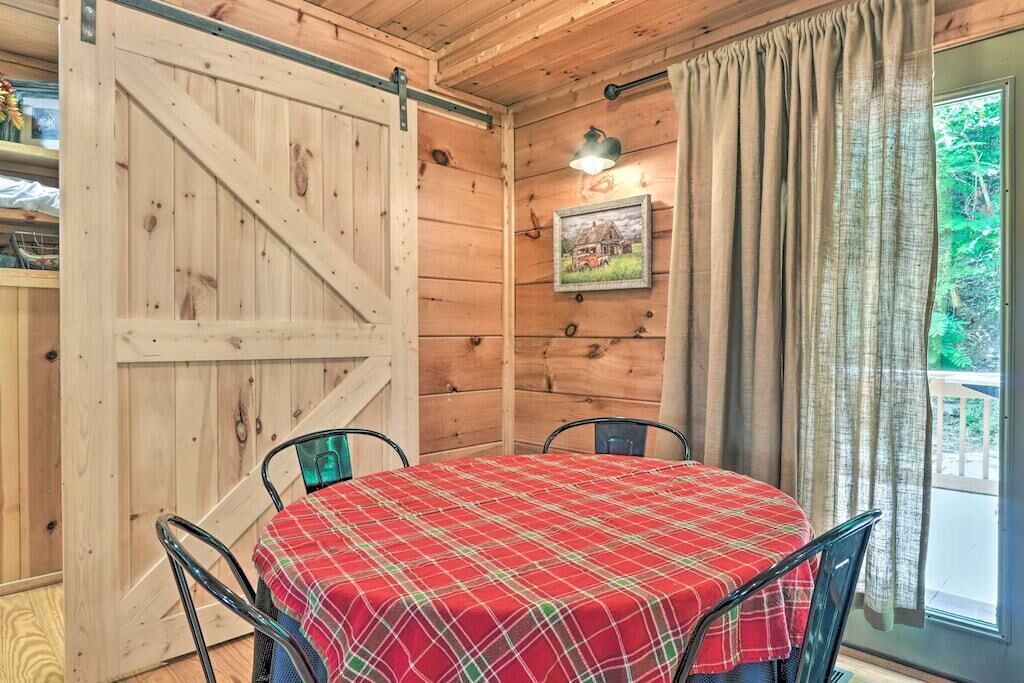 A wooden interior with a dining table covered in a plaid tablecloth, metal chairs, a sliding barn door, a framed picture on the wall, and a window with curtains.