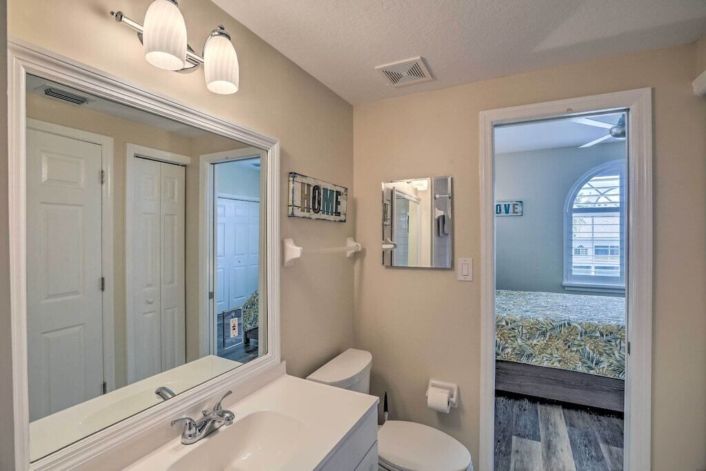 A bathroom with a large mirror, white fixtures, a toilet, and a view of a bedroom with a bed and a window.