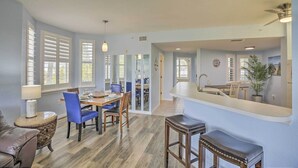 Een ruime eethoek met een houten tafel en blauwe stoelen, een keuken met een groot schiereiland, en een woonkamer met een leren bank en een bijzettafeltje.