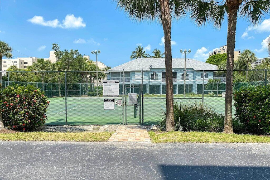 Zona de canchas de tenis delimitada por una valla, con un edificio al fondo, rodeada de palmeras y vegetación.