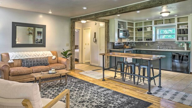 En moderne stue med en lædersofa i brunt, et træbord og et tæppe med mønster. I baggrunden ses et køkkenområde med en barstol og et spisebord.