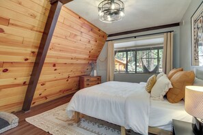 Ein Schlafzimmer mit holzverkleideter Schräge, einem Bett mit weißer Bettwäsche, einer Nachttischlampe und Blick ins Freie durch ein Fenster.