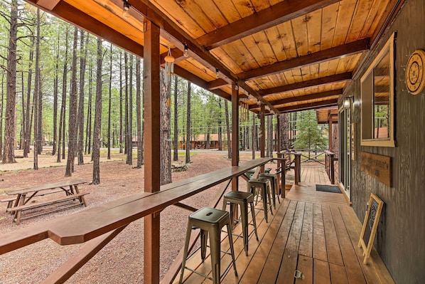 An outdoor wooden deck with picnic tables and bar stools, surrounded by a forest.