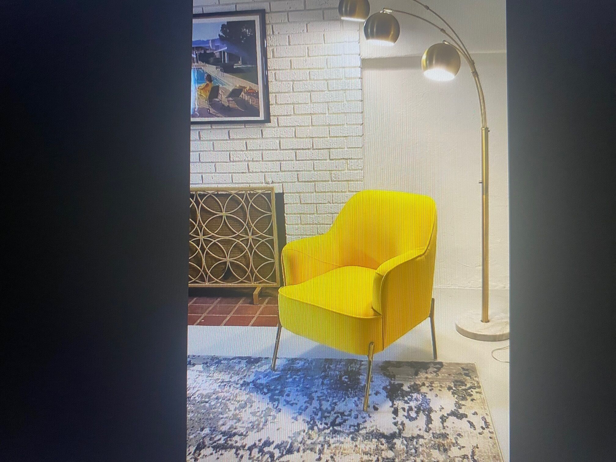 Ein Zimmer mit einem gelben Sessel, einer weißen Ziegelwand, einem gerahmten Bild, einem geometrisch gemusterten Bildschirm und einer Stehlampe.
