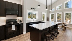 A modern kitchen with dark cabinetry, a large island with bar stools, and a view of the outdoors through large windows. - Air-Conditioning, Privacy, Family Retreat! (Durango)
