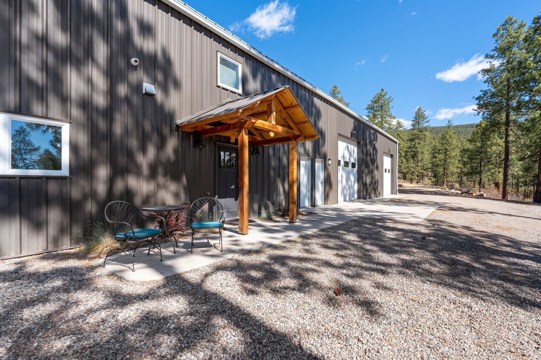 A modern building with a covered patio area, a gravel driveway, and a forested backdrop.
