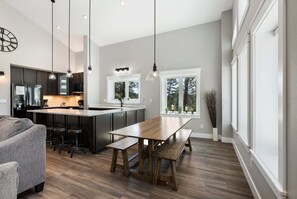 Una cocina moderna con una isla central, iluminación colgante y un amplio ventanal con vista a árboles.