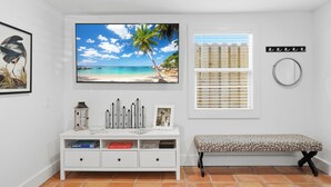 A room with a white console table, a bench, a large beach-themed TV, a window with blinds, and a wall-mounted mirror. - Canal-Front Oasis in Terra Ceia: Pool, Patio, Dock (Terra Ceia)