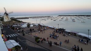 Un paseo marítimo con un molino, un puerto deportivo con barcos y una zona arenosa con gente y puestos de mercado.