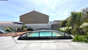 Una piscina con cerramiento de vidrio, sillas de descanso y un edificio grande al fondo.