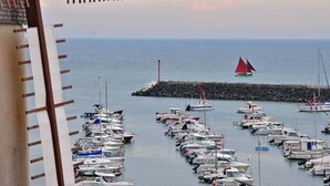 Una marina con numerosas embarcaciones amarradas, un velero a lo lejos y un rompeolas rocoso.