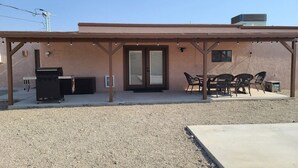 Un patio cubierto con una parrilla, mobiliario exterior y un edificio con puerta corrediza de vidrio.