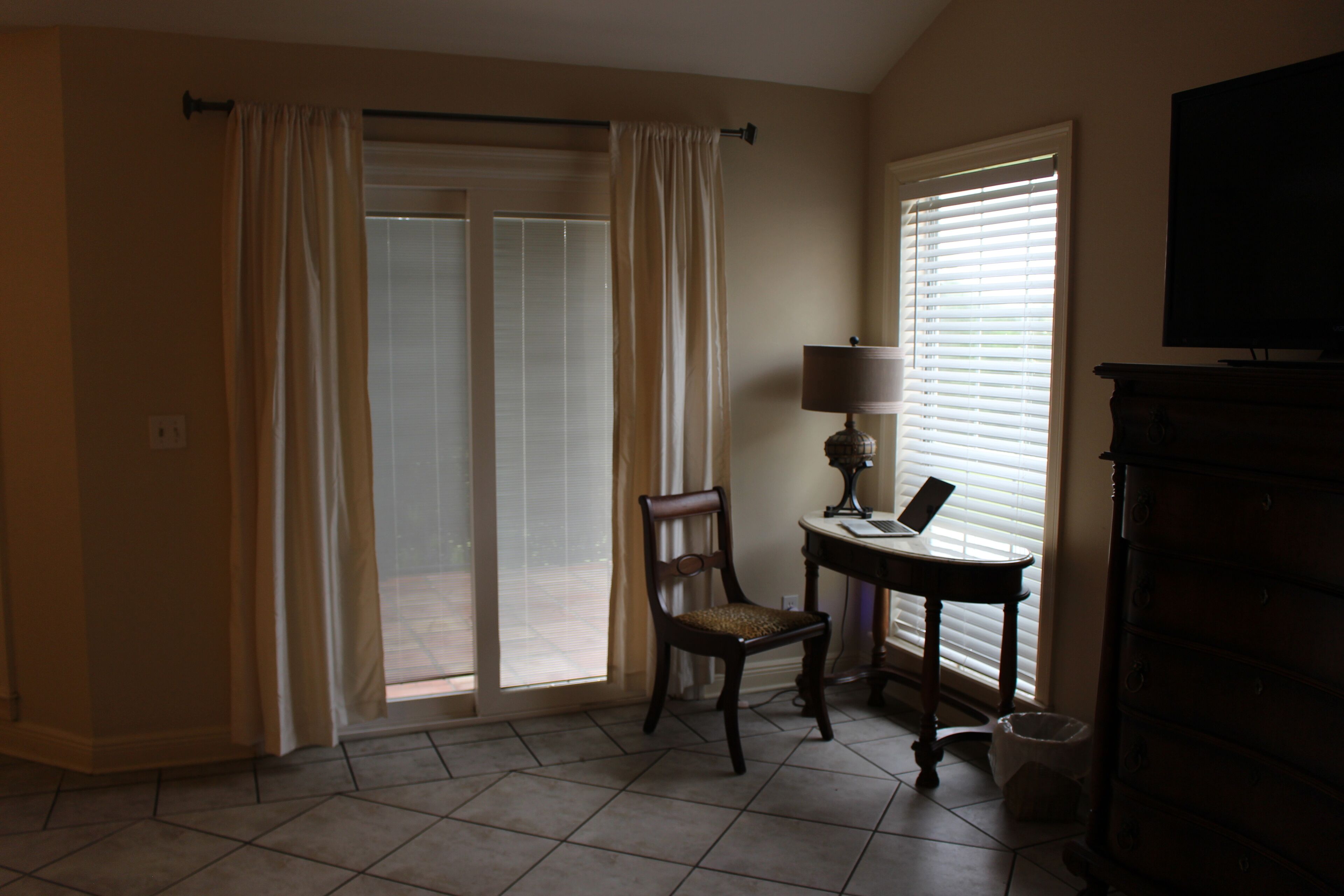 A room with a chair, a table with a lamp, a television, and a window with blinds.