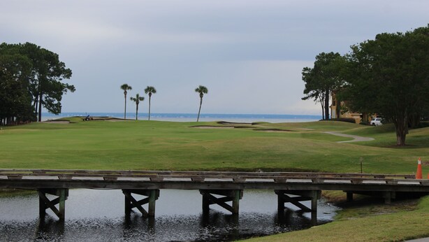 Un parcours de golf avec un cours d’eau, des palmiers et une vue lointaine sur l’océan.
