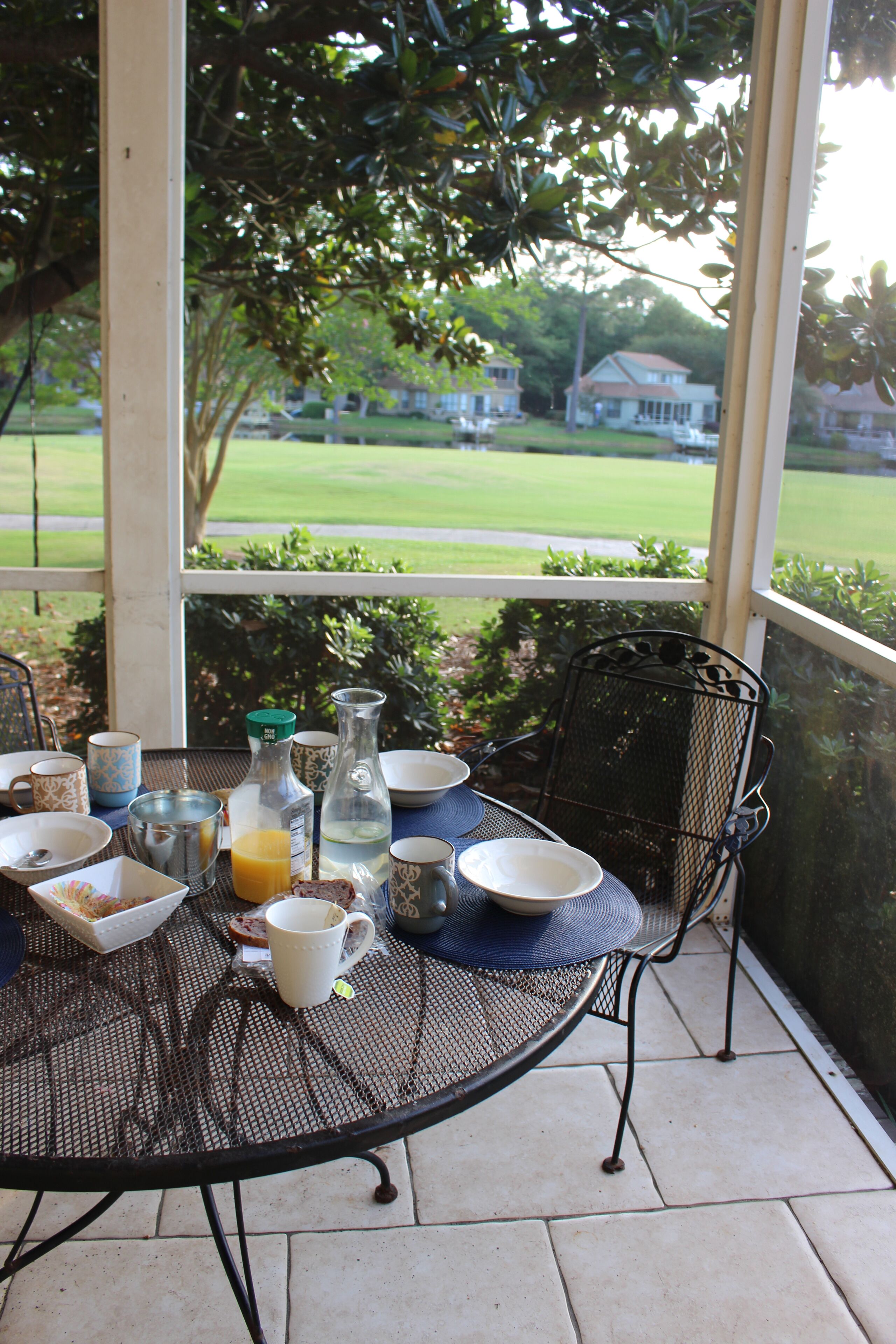 Une terrasse couverte avec une table mise pour le petit-déjeuner, comprenant des assiettes, des tasses et une carafe de jus d’orange.