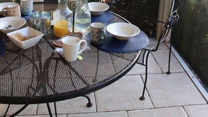 Une terrasse couverte avec une table mise pour le petit-déjeuner, comprenant des assiettes, des tasses et une carafe de jus d’orange.