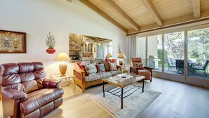 Un salon spacieux avec un fauteuil en cuir, un canapé et une table basse. Un mur en pierre et une grande fenêtre offrent une vue sur l’extérieur.