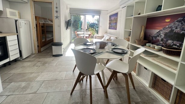 A compact kitchen with white cabinets, a dining area with white chairs, and a view of a garden through a glass door.