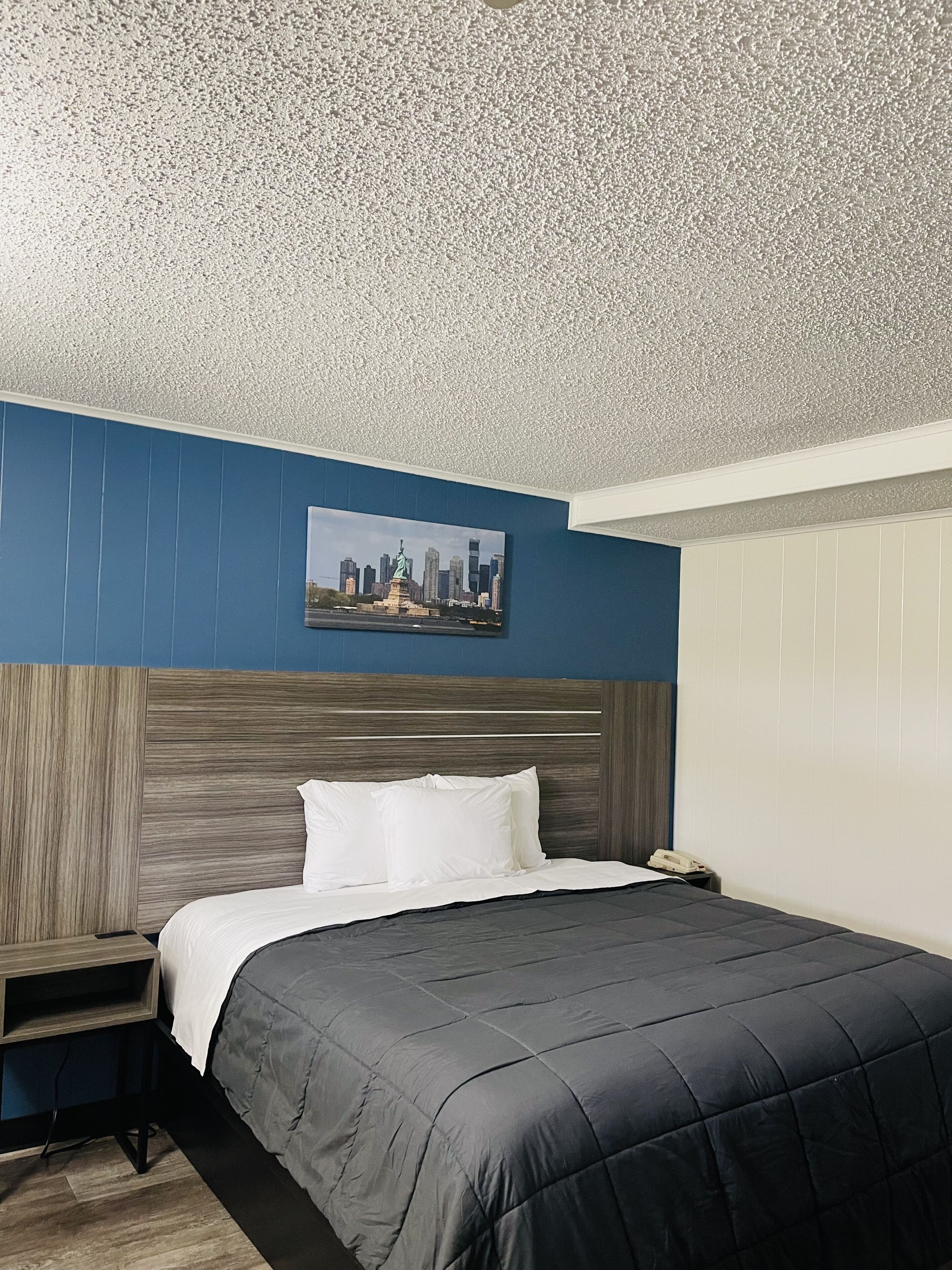 Een hotelkamer met een bed, nachtkastje, stadsgezicht schilderij en een gestructureerd plafond.