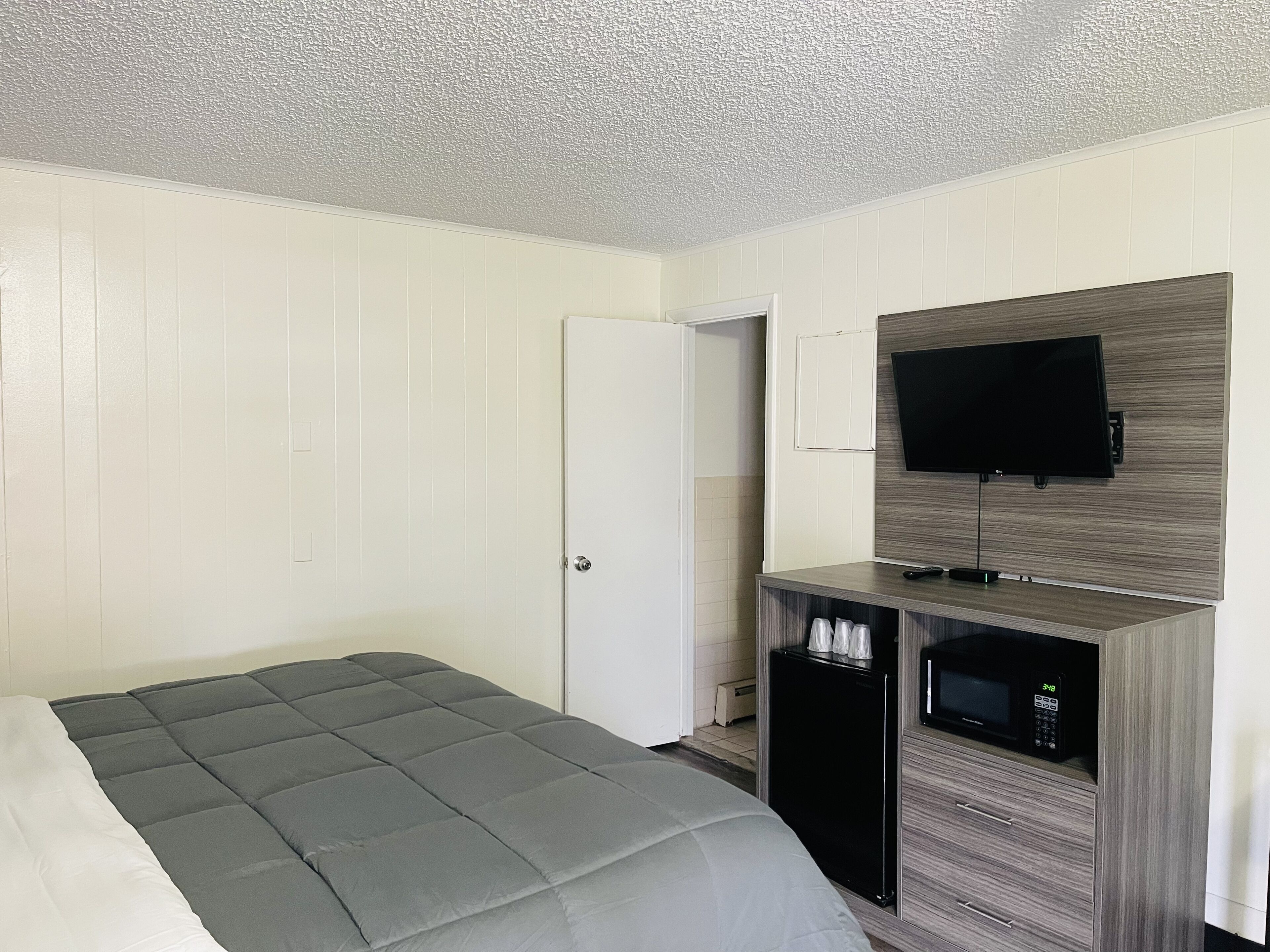 A hotel room with a bed, a TV mounted on a wooden panel wall, a small desk with a microwave, and a door leading to another room.