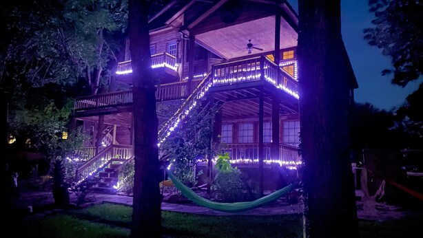 Een houten huis met twee verdiepingen, een balkon en een trap, omringd door bomen.