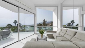 Ein modernes Wohnzimmer mit einer großen Eckcouch, einem runden Couchtisch und Blick auf Palmen sowie eine entfernte Landschaft durch raumhohe Fenster.