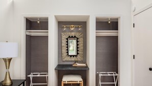 Une chambre à coucher avec un grand lit, un bureau avec une lampe, deux chaises, une coiffeuse avec un miroir et une fenêtre située au-dessus de la coiffeuse.