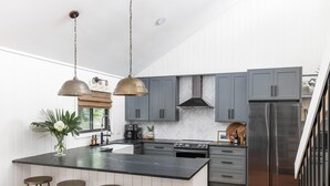 Une cuisine moderne avec des armoires grises, un comptoir noir et un réfrigérateur en acier inoxydable. On y trouve trois tabourets près de l’îlot central et un luminaire suspendu.