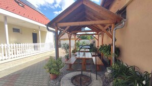 Un espacio exterior cubierto con mesas de picnic y una chimenea, rodeado de plantas en macetas y una valla blanca.