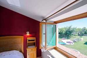 A bedroom with a wooden headboard, a small wooden nightstand, a bedside table with a lamp, and a large window with a view of a swimming pool and garden.