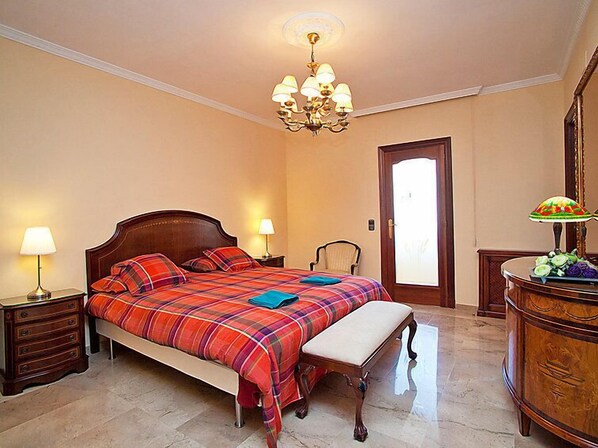 Une chambre à coucher avec un grand lit, deux tables de chevet avec des lampes, un banc en bois, une commode et un lustre.