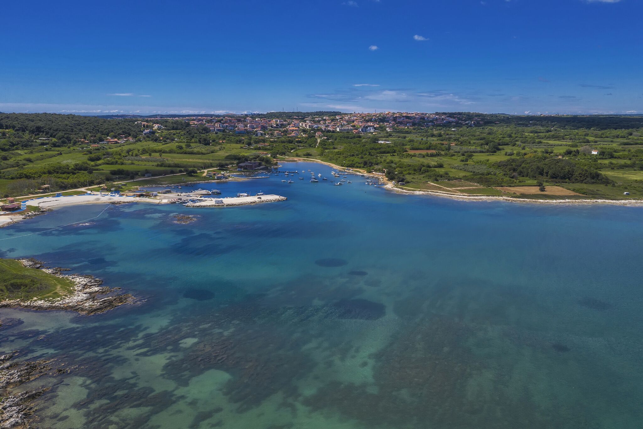 Vista aèria d'una zona costanera amb aigües blaves cristal·lines, una marina plena de vaixells i un poble al lluny.