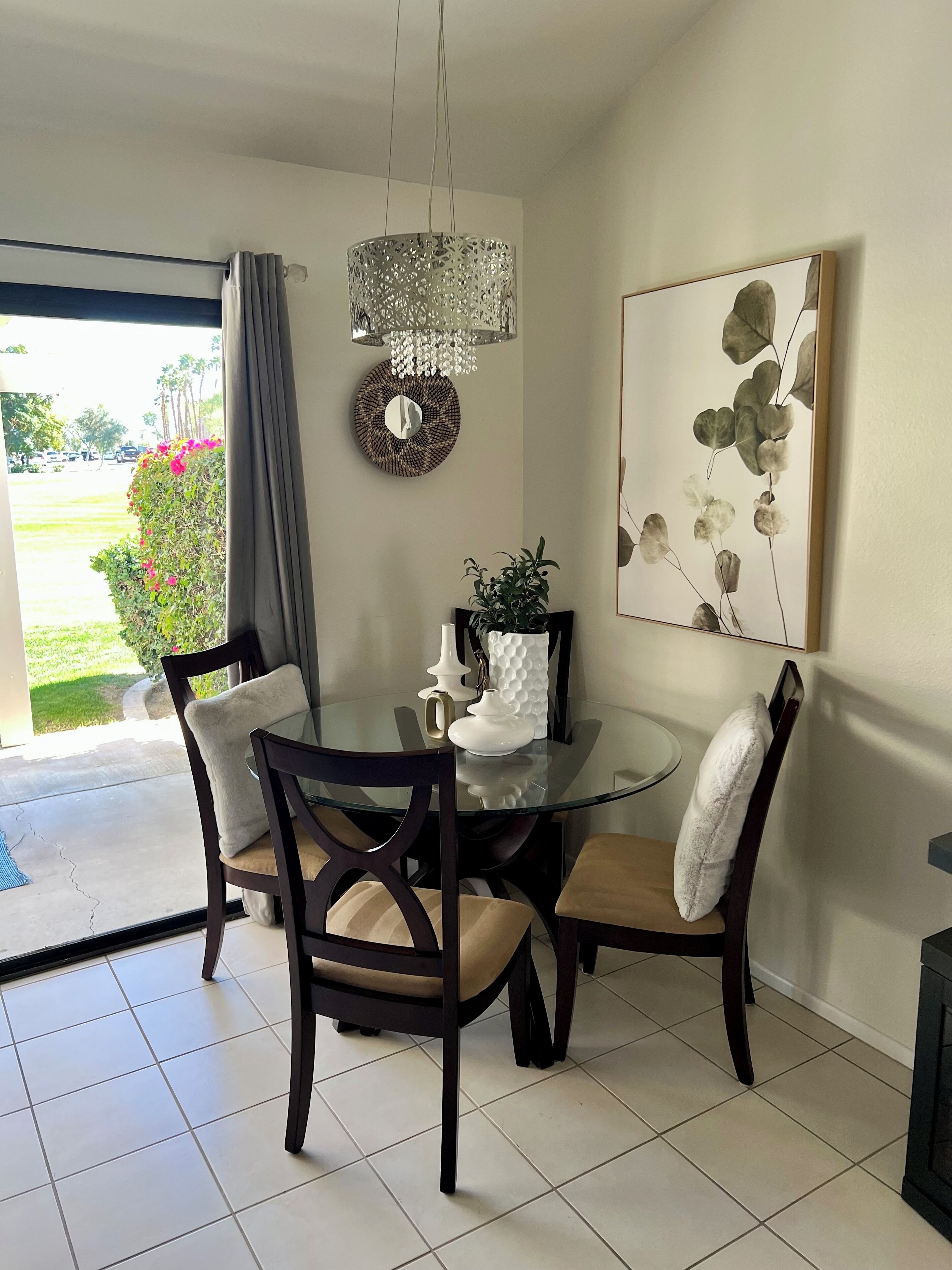 A dining area with a round glass table, four chairs, a chandelier, a wall-mounted mirror, and a framed botanical print.