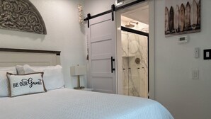 A neatly made bed with a pillow that reads 'BE OUR GUEST,' a wall-mounted clock, and a door leading to a bathroom with a shower.