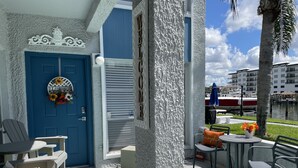 A coastal hotel entrance with a blue door, a welcome sign, outdoor seating, and a view of the marina.