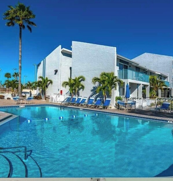 Une piscine entourée de chaises longues et de palmiers, le tout bordé d’un bâtiment moderne doté de balcons.