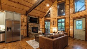 Ein Blockhaus-Innenraum mit Kamin aus Stein, einer modernen Küche mit Edelstahl-Kühlschrank und einer gemütlichen Sitzecke.