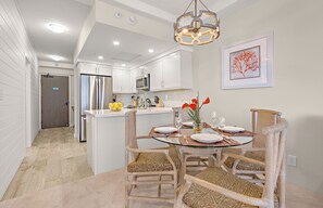 A modern kitchen with white cabinets, a stainless steel refrigerator, and a dining area with a glass table and wicker chairs.
