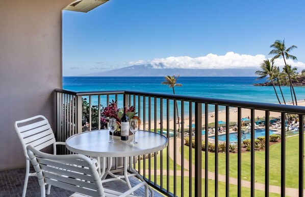 A balcony with a table and chairs, overlooking a beach with a pool and palm trees.