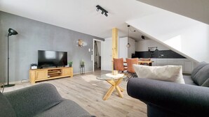 Ein modernes Wohnzimmer mit Flachbildfernseher, grauen Sofas, einem Holztisch und einem Essbereich mit Stühlen.
