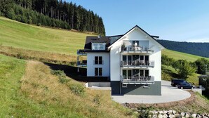 Ein zweistöckiges Haus mit Balkon, Auffahrt und einem bewaldeten Hügel im Hintergrund.