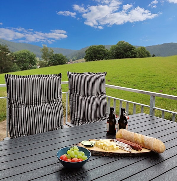 Une terrasse avec deux chaises à rayures, un saladier de fruits, une planche de fromages accompagnée de charcuteries et deux bouteilles de bière.