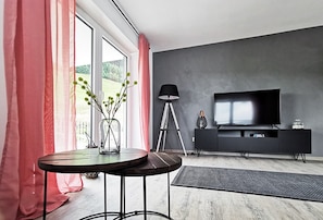 A modern living room with a large window, a round table, a tripod floor lamp, a flat-screen TV, and a dark-colored entertainment unit. - Apartment "Rehkitz" in Black Forest with Balcony & Wi-Fi (Baiersbronn)