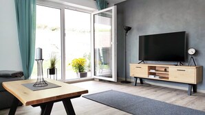 Una sala moderna con una mesa de madera, un televisor sobre un mueble bajo, una lámpara de pie y un ventanal corredizo que da a un espacio exterior con plantas.