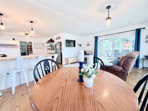 A modern kitchen with white cabinets, a dining table with chairs, and a living area with a sofa and a TV.